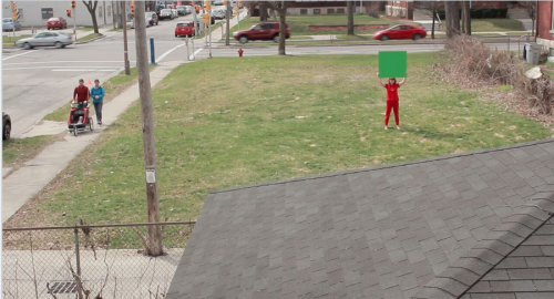 A scene from one of her videos, a performance in which Knapp revisits Milwaukee sites of mass protests of the killing of unarmed black man Dontre Hamilton. This time she is there alone, holding a sign that is a green screen, i.e. a sign on which passersby project their own thoughts and feelings.