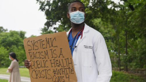 A doctor holds a sign about systemic racism and its relation to public health.