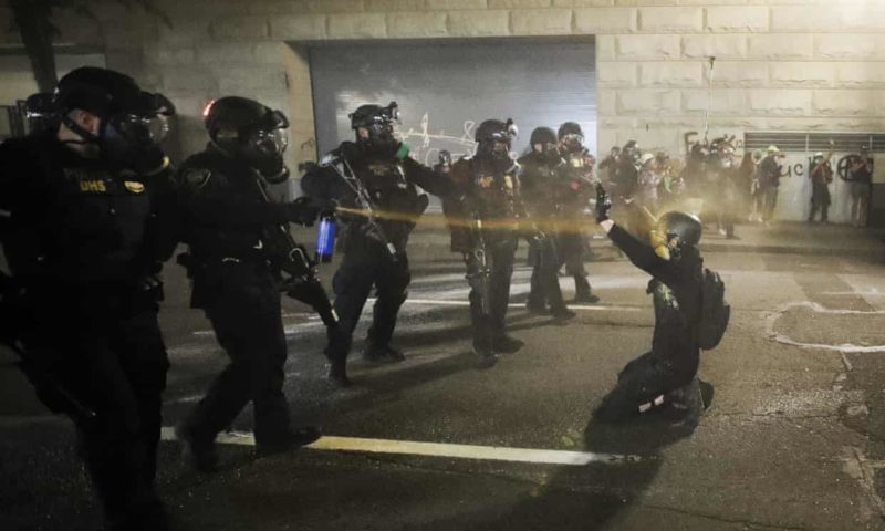 Police in riot gear spray tear gas in the face of a single kneeling protester