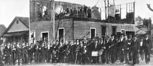 Black and white photo of white men with guns in front of a burned building that once was the Record newspaper building