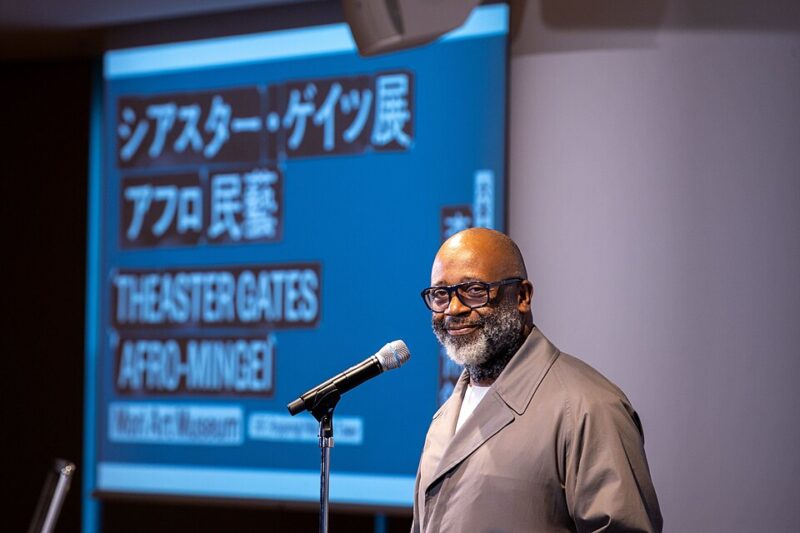 Theaster Gates stands near a microphone in front of a large screen